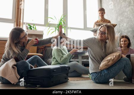 Sorridente studente energico dare alti cinque sul pavimento mentre imballare bagagli con roba nel soggiorno, mostrare fuori amico femminile Foto Stock