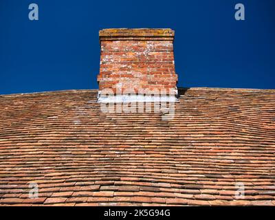 Irregolari, intemperie, vecchie piastrelle di argilla e camino su un tetto tradizionale cottage in Inghilterra, Regno Unito, preso in una giornata di sole con un cielo blu. Foto Stock