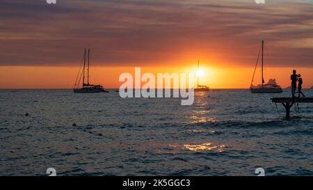 Silhouette di due persone che guardano il tramonto dal molo sul mare con barche a vela ancorate sull'isola di Formentera in Spagna Foto Stock