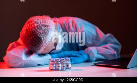 Covid-19 fatica. Ricerca sul vaccino. Insonnia pandemica. Medico donna stanco esausto in dpi che dorme sul posto di lavoro con fiale di dose per laptop in rosso blu n Foto Stock