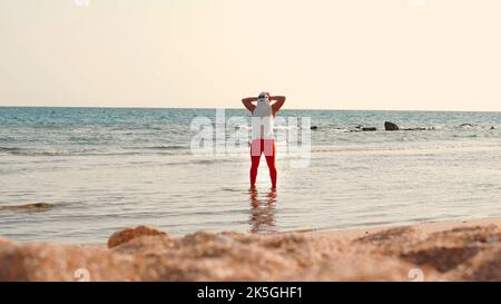 Babbo Natale si bagna il sole. Babbo Natale divertendosi. Babbo Natale divertente, in occhiali da sole, in piedi in acqua di mare sulla spiaggia. Vacanze estive a Babbo Natale. Foto di alta qualità Foto Stock