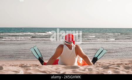 Vacanze estive a Babbo Natale. Babbo Natale divertendosi. Babbo Natale divertente, in pinne, rilassarsi mentre si siede sulla spiaggia di sabbia sul mare. Vista sul retro. Foto di alta qualità Foto Stock