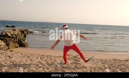 Vacanze estive a Babbo Natale. Babbo Natale divertendosi. Funny Babbo Natale, in pinne corre attraverso la spiaggia sabbiosa fino al mare. Ha fretta di nuotare, fare snorkeling. Vista posteriore. Foto di alta qualità Foto Stock