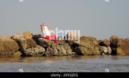 Babbo Natale si bagna il sole. Babbo Natale divertendosi. Babbo Natale divertente, in occhiali da sole e pinne seduti sulla spiaggia rocciosa sul mare. Vacanze estive a Babbo Natale. Foto di alta qualità Foto Stock