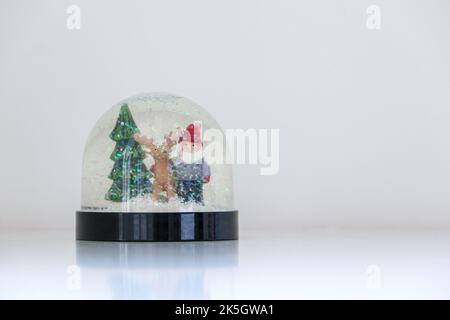 Un globo di neve di Natale scintillante e scintillante con la neve che cade su Babbo Natale, una renna, e un albero di Natale, su sfondo bianco innevato, copia sp Foto Stock