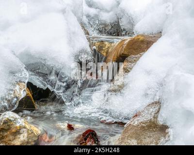 Ruscello di montagna che scorre sotto la neve bianca attraverso le rocce e le tartarughe di ghiaccio. Foto Stock