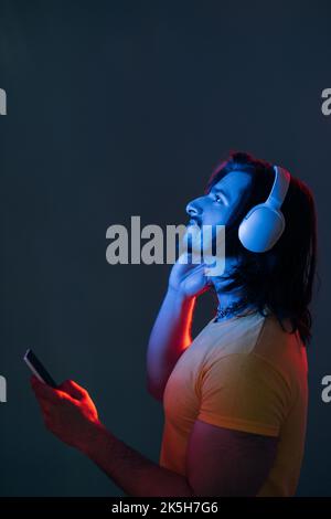 Musica di tendenza. Uomo Hippster. Ritratto al neon. Tecnologia mobile. Un ragazzo ispirato alla beared in cuffie wireless che tengono lo smartphone alla ricerca di spazio copia Foto Stock
