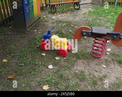 Moto di plastica colorata e un cavallo di legno giocattolo a dondolo in un parco giochi con una panca di legno, recinzione di legno e cestino sullo sfondo Foto Stock