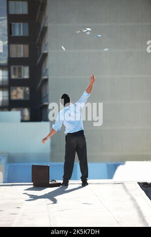 La sensazione di lasciar andare le mie catene finanziarie. Un uomo d'affari in piedi sul bordo di un tetto che getta soldi nell'aria con copyspace. Foto Stock