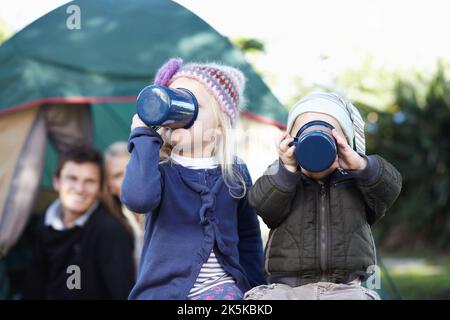 Preparandosi per un campeggio di giorni energici. Genitori amorosi che guardano i loro bambini bere da tazze mentre fuori campeggio. Foto Stock