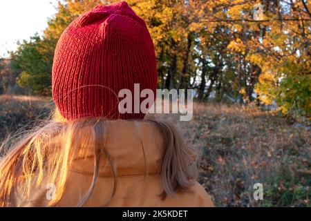 Giovane donna con lunghi capelli biondi in un cappello rosso e cappotto giallo camminando nel parco autunnale. Vista posteriore. Colori autunnali brillanti. Tempo soleggiato. Godetevi la vita Foto Stock