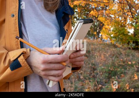 Donna in giacca gialla con un libro di disegno e una matita su sfondo giallo alberi autunnali nel parco. Primo piano delle mani con notebook e matita. Disegno in Foto Stock