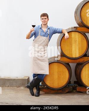 Degustare un buon vino rosso. Un giovane enologo che tiene un bicchiere di vino rosso mentre si appoggia su un barile. Foto Stock