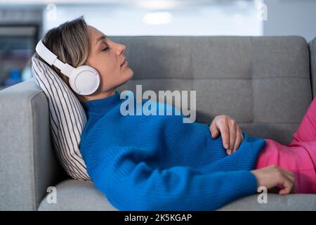 Una giovane donna che ascolta la musica con le cuffie wireless chiude gli occhi e si appoggia sul comodo divano Foto Stock