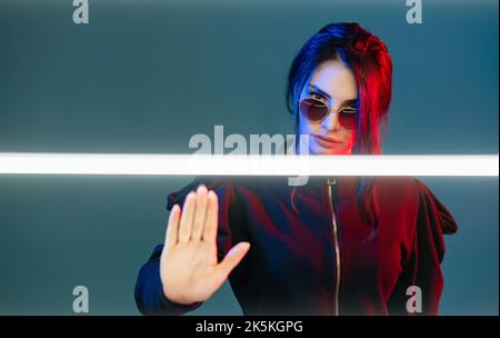 Basta con la discriminazione. Ritratto al neon. Empowerment femminista. Donna severa in occhiali da sole in rosso blu bagliore con LED bianco avvertimento con nope gesto i Foto Stock