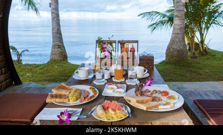 Tavolo per la colazione sulla spiaggia con palme in Thailandia. Colazione colorata con uova e frutta Foto Stock