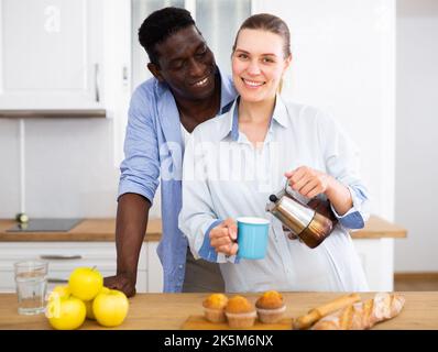 Felice coppia biraciale ridendo e bevendo caffè la mattina in cucina Foto Stock