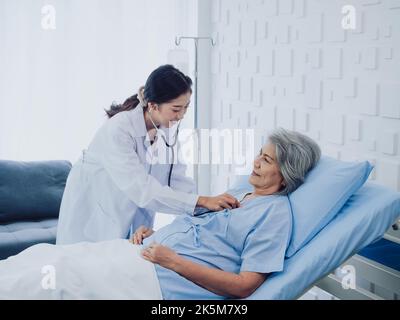 Una bella giovane donna asiatica medico in vestito bianco sorrise mentre utilizzando stetoscopio per esaminare, ascoltare il battito cardiaco di anziana paziente femminile in blu Foto Stock