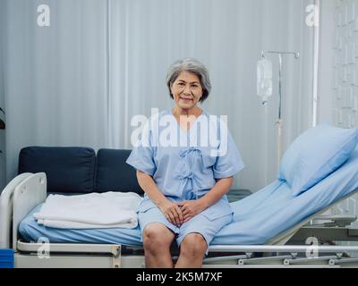 Felice sorriso bella asiatica anziana paziente con chirurgia del ginocchio in vestito azzurro seduto sul letto vicino al palo medico IV stand con soluzione salina Foto Stock