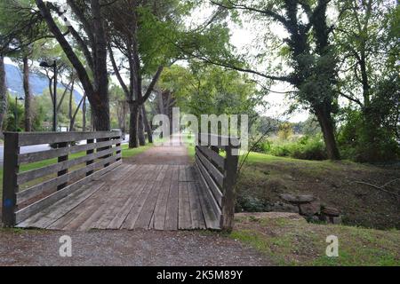 Vista sul verde del grande marciapiede di sabbia nel verde del parco cittadino con ponte in legno sul torrente di montagna. Fa parte della pista ciclabile Lecco-Milano. Foto Stock