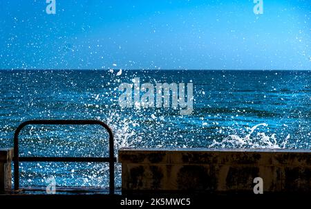 Le onde blu del mare si schiantano contro il molo creando un'esplosione di schiuma bianca in un chiaro pomeriggio di sole Foto Stock