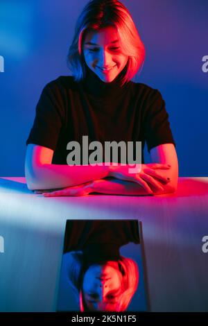 Disturbo bipolare. Ritratto al neon. Cambiamento di umore. Felice ragazza sorridente con triste depresso specchio viso riflesso in rosso blu luce isolato al buio. Foto Stock