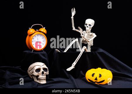 Halloween. Un giocattolo Skeleton che striscia fuori dalla tomba, e agitando la mano, saluta, zucche con le grimaces, un orologio arancione, un cranio su un bac nero Foto Stock