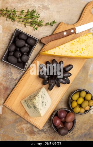 Due pezzi di formaggio e coltello sul tavolo. Olive verdi, rosse e nere in ciotole. Rametto di timo e grappolo di uva nera. Giacitura piatta. Metallo arrugginito b Foto Stock