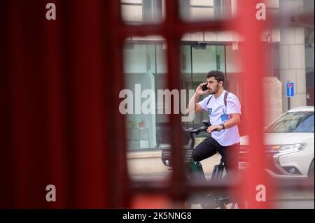 LONDRA - 21 maggio 2022: L'uomo usa il telefono cellulare mentre noleggiasse una bicicletta elettrica. Vista tramite la tradizionale cassetta telefonica Foto Stock