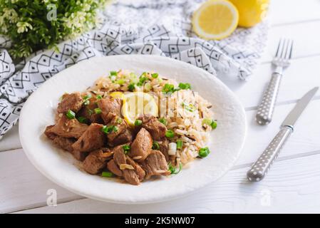 Grossi pezzi di maiale accanto al riso bollito a grani lunghi, cosparsi di cipolle verdi tritate e una fetta di limone. Un appetitoso servizio di pranzo sostanzioso Foto Stock