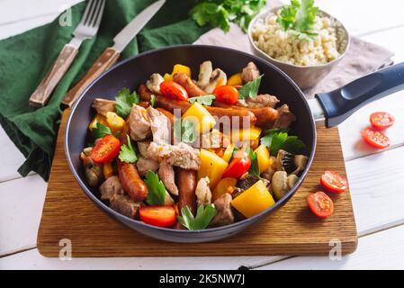 Salsicce fritte, grossi pezzi di carne di maiale, con peperoni gialli, fette di pomodoro ciliegia, foglie di prezzemolo e couscous. Un piatto colorato in una padella. Foto Stock