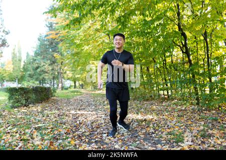 Uomo al mattino Jog nel parco autunnale, sportivo asiatico in abbigliamento sportivo con cuffie e telefono in foresta ascolta la musica su Jog e camminare stile di vita attivo Foto Stock