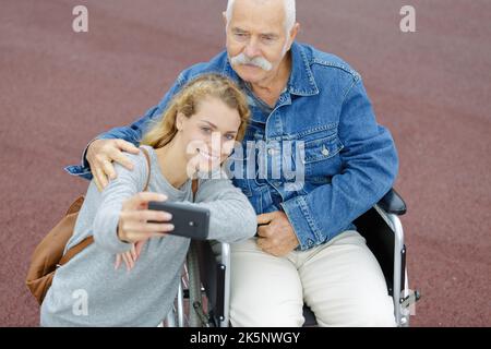 uomo anziano in sedia a rotelle e giovane donna che prende selfie Foto Stock