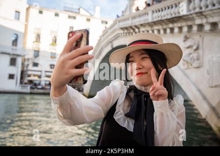 Sorridente turista fa il segno V di vittoria sotto il ponte di Rialto a Venezia mentre prende un selfie. Foto Stock