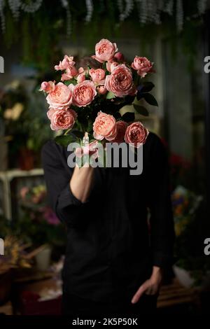 Uomo in camicia nera con bouquet di fiori. Cute maschio caucasico che tiene un mazzo di rose rosa fresche di fronte al suo volto che lo nasconde. Immagine tasto bassa. Concetto di amore o romanticismo. Foto di alta qualità Foto Stock