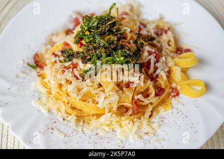 Tagliatelle italiane bollite con pancetta a cubetti fritta, formaggio grattugiato e rucola fritta su piatto bianco, primo piano, ristorante all'aperto. Foto Stock