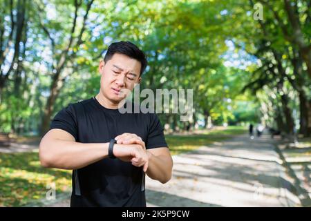 un atleta maschile infelice e sconvolto che guarda al fitness con lo smartwatch, il risultato di un allenamento cattivo, l'uomo asiatico che si esercita nel parco autunnale di giorno. Foto Stock