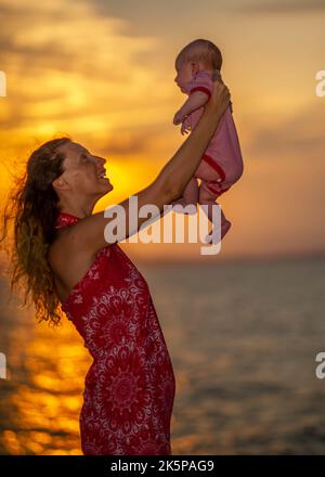 Madre che solleva il bambino in aria sopra il cielo panoramico tramonto indossando vicino rosso Foto Stock