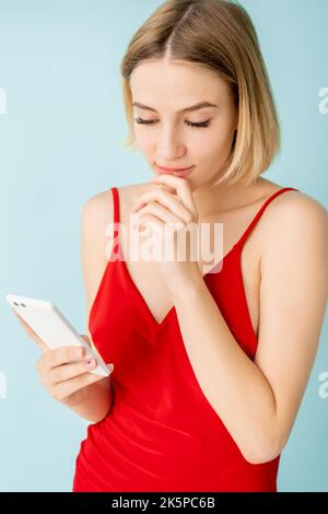 Donna dubbiosa. Comunicazione mobile. Amicizia virtuale. Bella donna premurosa che tiene lo smartphone sensazione di esitazione isolato blu. Foto Stock