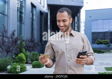 Un giovane imprenditore effettua acquisti online, utilizza uno smartphone e una carta di credito bancaria, un uomo fuori da un ufficio cammina per la città durante una pausa al lavoro. Foto Stock