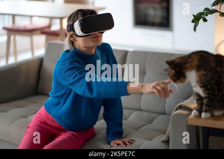 Giovane donna in occhiali VR esplora il gioco e cerca di toccare il gatto domestico seduto sul tavolo di legno Foto Stock
