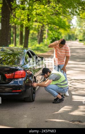 Addetto alla manutenzione che esamina la ruota rotta di un'auto rotta nella strada forestale Foto Stock