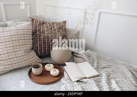 Colazione a letto, tavolo da gioco con sedie e divano. Tazza di caffè, zucche. Apri libro. Tazza di caffè sul vassoio. Asciugare il bouquet di erba nel vaso a forma di sfera Foto Stock