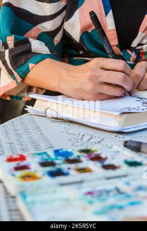 foto verticale di mani caucasiche di una donna irriconoscibile seduta all'aperto facendo un disegno con pennarello e carta, con acquerelli sfocati nella f Foto Stock