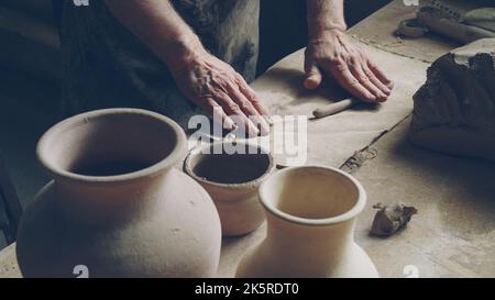 Mani sporche di ceramista maschio senior stanno rotolando piccolo pezzo di argilla su grande tavolo di lavoro. Vasi e vasetti in ceramica fatti a mano, utensili per vasaio sono visibili. Foto Stock