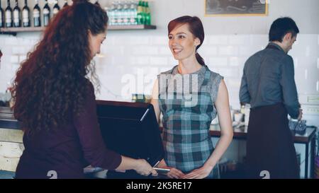 I lavoratori allegri di un'accogliente caffetteria salutano i clienti, prendono i pagamenti con lo smartphone utilizzando il terminale delle carte di credito e vendono bevande da asporto ai giovani. Foto Stock