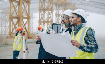 L'architetto uomo in tuta parla con un ingegnere nell'area degli edifici, mentre i topografi lavorano con attrezzature moderne in background. Concetto di persone e affari. Foto Stock