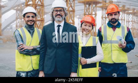 Il team multirazziale di architetti di successo sta in piedi all'aperto nel sito del progetto sorridendo tenendo documenti e progetti. Persone e concetto di occupazione. Foto Stock