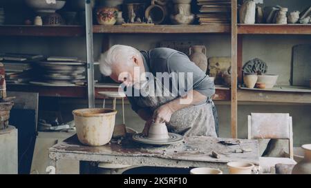 Il vasaio professionale dell'uomo bearded anziano sta facendo la ciotola dall'argilla sulla ruota del vasaio rotante, allargando e livellando il clayware bagnato. Diverse figure in ceramica fatte a mano sullo sfondo. Foto Stock