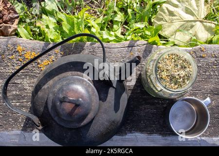 Vecchio bollitore di ferro d'annata, vaso con erbe e tazza di tè alle erbe fresco su sfondo di legno. Preparazione di tè alle erbe sano all'aperto. Foto Stock